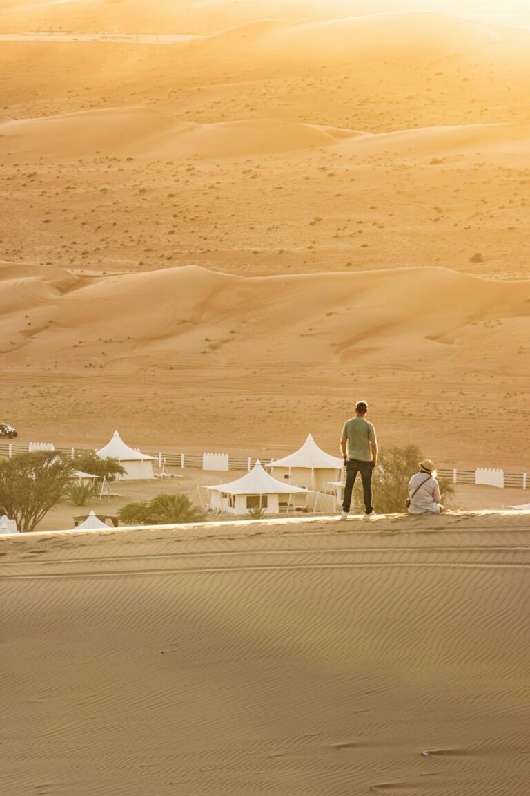 Travelers watching a desert camp from the top of a golden sand dune